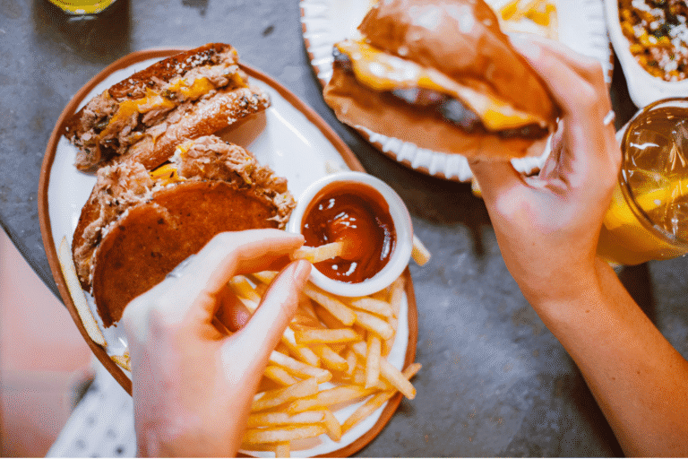 Osoba siedząca przy stole trzyma burgera i sięga frytką do miseczki z keczupem. Na talerzu leżą drugi burger i frytki, a obok stoją napoje i dodatkowe porcje jedzenia.