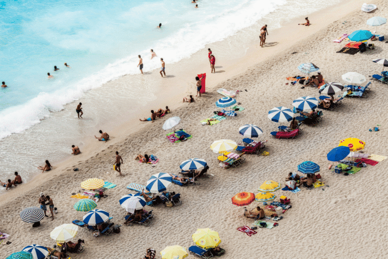 Widok z góry na zatłoczoną plażę. Na piasku i w wodzie wypoczywa wiele osób, wokół rozstawione są kolorowe parasole i leżaki, a przy brzegu widać fale rozbijające się o plażę.