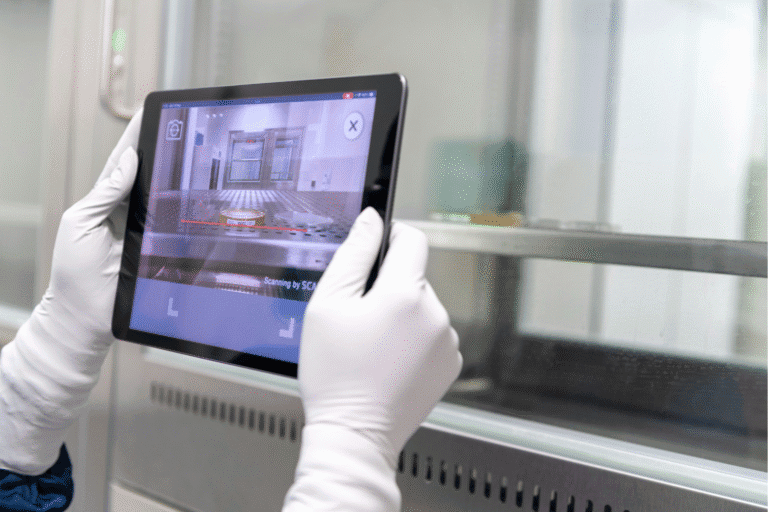 Osoba w rękawiczkach ochronnych trzyma tablet, na którym wyświetlany jest obraz wnętrza laboratoryjnego urządzenia oraz dane z procesu monitorowania. Zdjęcie wykonano w laboratorium lub zakładzie badawczym.