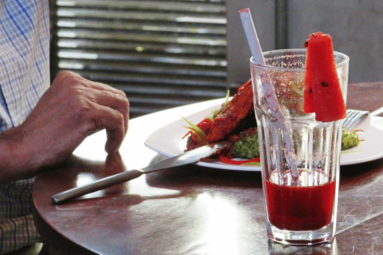 Szklanka czerwonego napoju z kawałkiem arbuza na stole w restauracji, obok osoba jedząca posiłek.