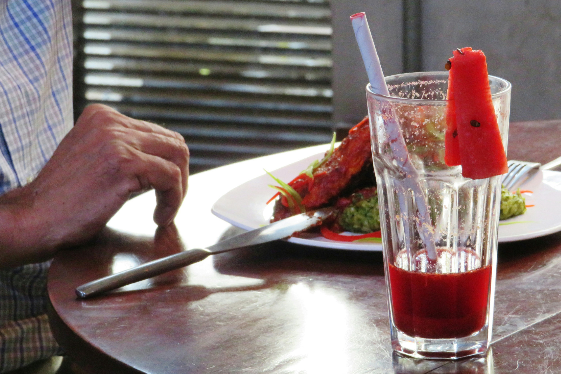 Szklanka czerwonego napoju z kawałkiem arbuza na stole w restauracji, obok osoba jedząca posiłek.